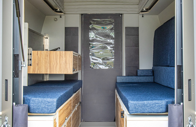 mid Groß-Gerau - Blick durch die geöffnete Heckklappe in den Innenraum des PlugVan Wohnmoduls im Nissan Interstar mit braunen Schränken und blauen Sitzkissen. Nissan